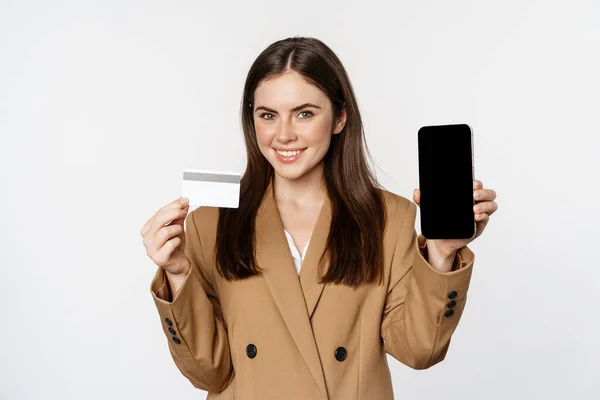 Mujer de negocios mostrando la pantalla del teléfono móvil y la tarjeta de crédito, demostrando aplicación en el teléfono inteligente, de pie en traje sobre fondo blanco — Foto de Stock