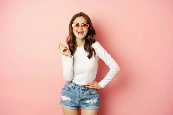 Portrait of stylish glam girl in sunglasses, laughing and smiling, standing over holiday pink background — Fotografia de Stock
