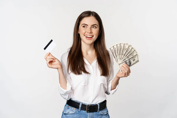 Gelukkige vrouw met geld en creditcard met doordacht gezicht, concept van lening en microkrediet, staande over witte achtergrond — Stockfoto