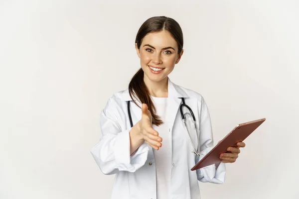 Friendly young female doctor, physician stretch out hand for handshake, greeting patient in clinic, holding digital tablet with medical data, white background — Foto de Stock
