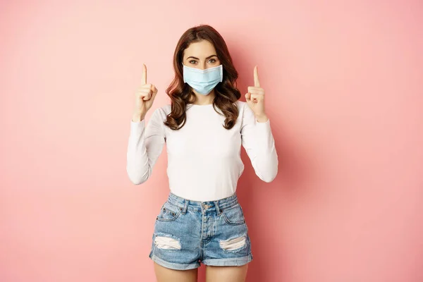 Portrait of young stylish woman in casual summer clothes, wearing face medical mask, pointing fingers up, showing promo offer, standing against pink background — Fotografia de Stock