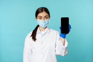 Young woman doctor in medical face mask and hospital uniform, showing mobile phone app, screen interface, online clinic concept, standing over torquoise background