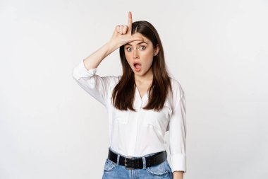 Young woman mocking loser, showing L word sign on forehead and gasping with disbelief surprise, standing against white background