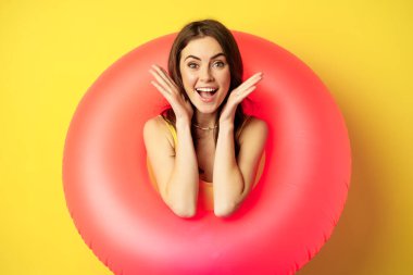 Happy beautiful young woman relaxing on summer vacation, wearing pink swim ring for beach holiday, standing against yellow background