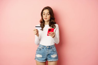 Online shopping. Young woman order online, making purchase in app, holding smartphone and credit card, standing against pink background