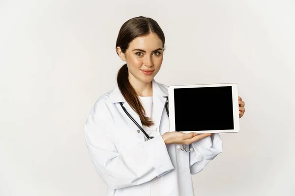 Ayuda médica en línea, cita remota. Sonriente hermosa mujer médico mostrando la pantalla de la tableta digital, demostrando el logotipo del sitio web, de pie sobre el fondo blanco — Foto de Stock