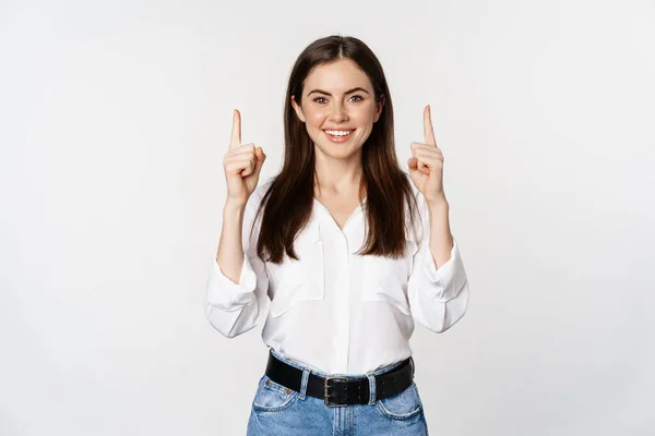 Sorrindo mulher bonita confiante, apontando os dedos para cima, mostrando banner logotipo, anúncio, de pé em blusa branca contra fundo estúdio — Fotografia de Stock