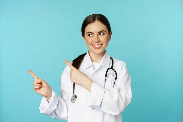 Portrait of smiling medical worker, girl doctor in white coat with stethoscope, pointing fingers left, showing medical clinic advertisement, torquoise background — Fotografia de Stock