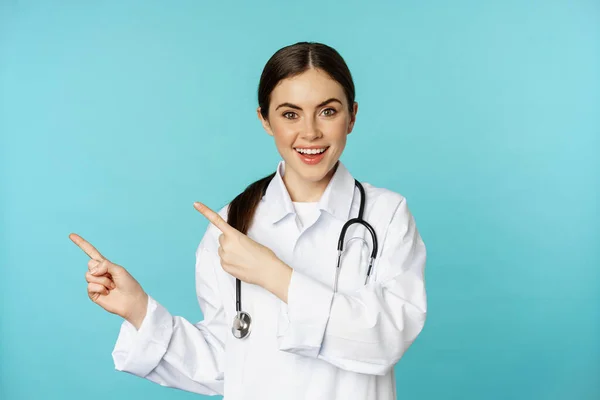 Gülümseyen sağlık çalışanı portresi, steteskoplu beyaz önlüklü kız doktor, solu işaret eden, tıbbi klinik reklamını gösteren torkuaz arka plan. — Stok fotoğraf
