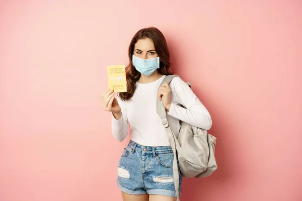Fiatal nő megy kirándulni, orvosi maszkot visel, mutatja kovid tanúsítvány, útlevél és aiplance jegyek, nyaralás világjárvány idején, rózsaszín háttér — Stock Fotó
