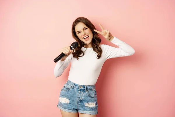 Party girl. Happy young woman singing in microphone, performing song, having fun at event, standing over pink background — Stockfoto