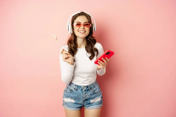 Stylish brunette woman dancing in headphones with mobile phone, listening music in earphones, licking lolipop, standing over pink background — Stockfoto