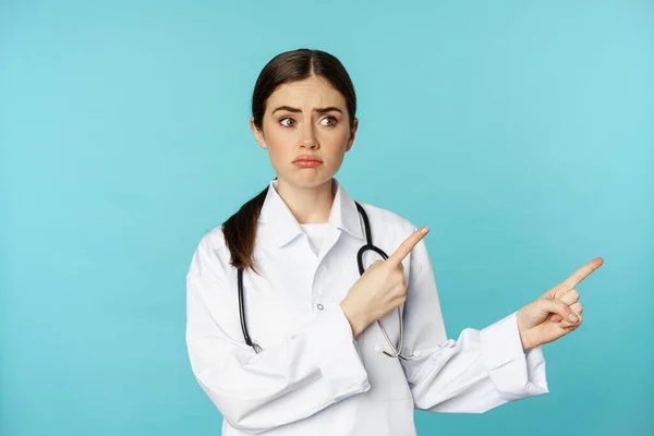 Retrato de médico decepcionado, mujer trabajadora médica señalando con los dedos hacia la derecha y mirando triste, arrepentimiento, expresión de la cara sombría, fondo azul — Foto de Stock