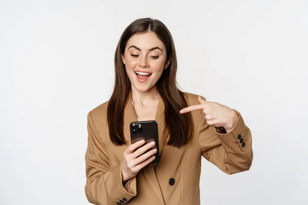 Enthusiastic saleswoman, business woman pointing finger at mobile phone and smiling, showing on cellphone, standing against white background — Stockfoto