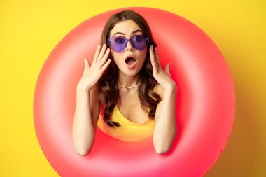 Portrait of young pretty woman in sunglasses, wearing beach swimming ring, looking surprised and amazed, shocked wow face, yellow background
