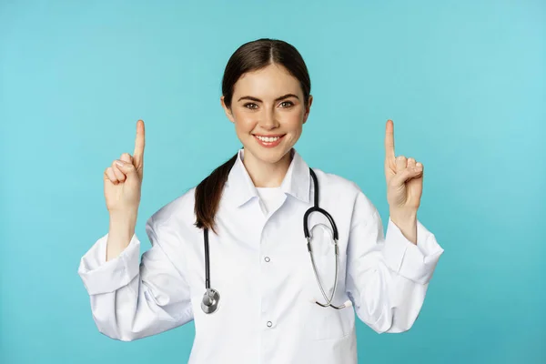 Porträt einer selbstbewussten jungen Ärztin, Ärztin im Mantel, mit erhobenen Fingern lächelnd, zeigt Medikamentenwerbung, Klinikwerbung, Haubenhintergrund — Stockfoto