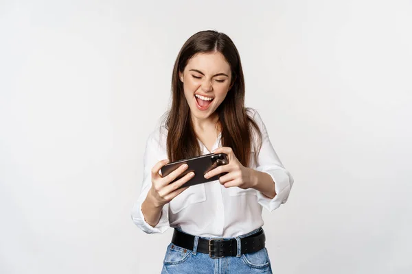 Mulher morena feliz jogando videogame móvel, sorrindo e olhando para a tela animada, em pé sobre o fundo branco — Fotografia de Stock