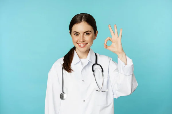 Portrait of satisfied, smiling medical worker, woman doctor showing okay, ok, zero no problem gesture, excellent sign, standing pleased over torquoise background — ストック写真