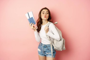 Beautiful girl on vacation, going on trip with passport and tickets, holding backpack. Young woman traveller backpacking, standing over pink background
