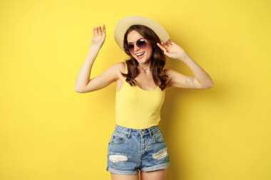 Stylish girl in straw hat and tank top, ready for summer, going on vacation and smiling pleased, standing over yellow background