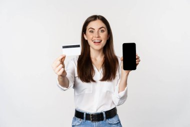Portrait of female model showing credit card with smartphone screen, recommending application, standing over white background
