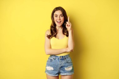 Creative young woman has suggestion, raising finger, eureka sign, has an idea, standing in summer outfit over yellow background
