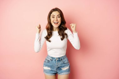 She is a winner. Enthusiastic brunette girl celebrating, jumping from happiness and shouting joyful, achieve goal success, standing over pink background
