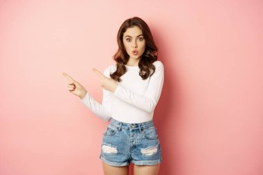 Wow, look there. Amazed feminine woman pointing fingers left, looking surprised and impressed, standing over pink background