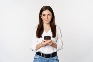 Akıllı telefonlu genç bir kadın, gülümsüyor ve kameraya bakıyor, cep telefonu uygulaması, cep telefonu teknolojisi ve online alışveriş konsepti kullanıyor, beyaz arka plan
