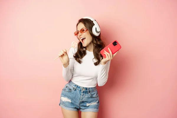 Summer girl laughing, eathing lolipop and listening music in headphones, dancing with smartphone against pink background — Zdjęcie stockowe