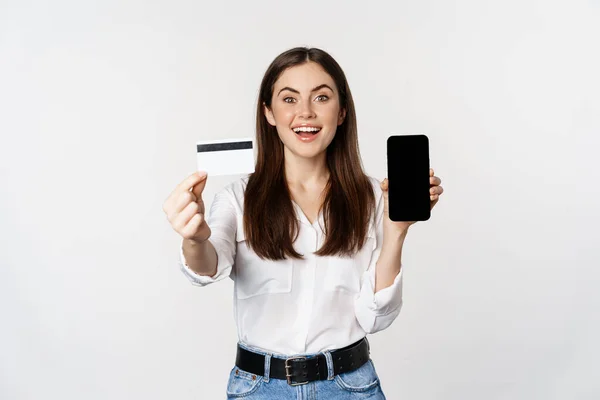 Retrato de modelo femenino mostrando tarjeta de crédito con pantalla de teléfono inteligente, recomendando la aplicación, de pie sobre fondo blanco — Foto de Stock