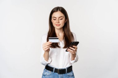 Portrait of young woman looking at smartphone screen, paying for purchase online with credit card, standing over white background, placing order