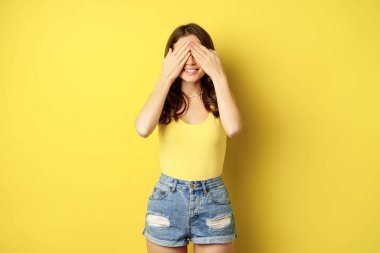 Portrait of brunette woman cover eyes with hands, blindfolded, smiling with anticipation, expecting smth, waiting for surprise, standing over yellow background
