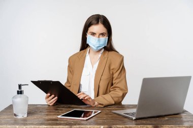 Covid, pandemic and office workplace concept. Portrait of corporate woman in medical face mask from coronavirus, working, standing over white background