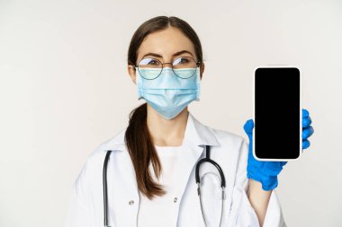 Çevrimiçi tıbbi yardım konsepti. Gözlüklü ve yüz maskeli kadın doktor, cep telefonu ekranını gösteriyor, hastalar için uygulama arayüzü veya web sitesi, beyaz arka planda duruyor