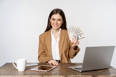 Portrait of businesswoman sitting in office with money, working and making profit income, posing happy against white studio background
