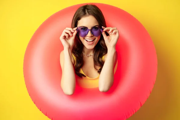 Close up portret stylowej młodej kobiety, dziewczyna z pierścieniem kąpielowym i okulary, gotowy na wakacje na plaży, wakacje, stojąc na żółtym tle — Zdjęcie stockowe