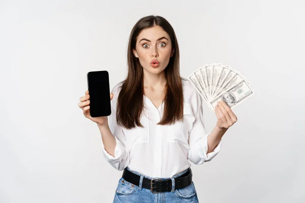 Mujer joven entusiasta ganar dinero, mostrando interfaz de la aplicación del teléfono inteligente y dinero en efectivo, microcrédito, concepto de premio, de pie sobre fondo blanco — Foto de Stock