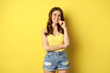 Thinking woman smiling, looking up and deciding, imaging smth, picturing or daydreaming, posing in summer clothes against yellow background