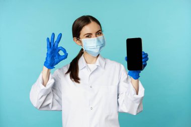 Portrait of doctor in medical face mask and gloves, showing mobile phone app, smartphone screen and okay sign, recommending online checkup website, standing over torquoise background