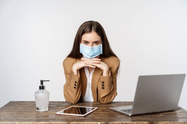 Workplace and covid-19 concept. Image of corporate woman at office, sitting at table with hand sanitizer and laptop, wearing face mask to protect from coronavirus