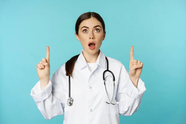 Hevesli bir tıp çalışanı, beyaz önlüklü genç bir kadın doktor, steteskop, reklam gösteriyor, parmakları havada, torkuaz arka planda duruyor. — Stok fotoğraf