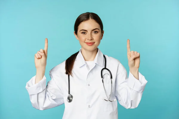 Portrait of confident young woman doctor, medical worker in coat, pointing fingers up and smiling, showing medicine advertisement, clinic promo, toquoise background — Stok fotoğraf
