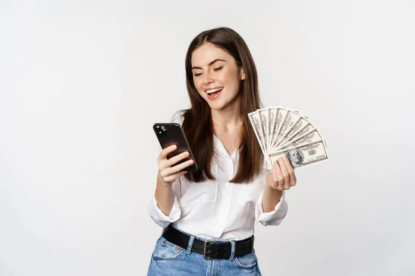 Retrato de mujer sonriente feliz usando aplicación de teléfono móvil, sosteniendo dinero en efectivo, de pie sobre fondo blanco —  Fotos de Stock