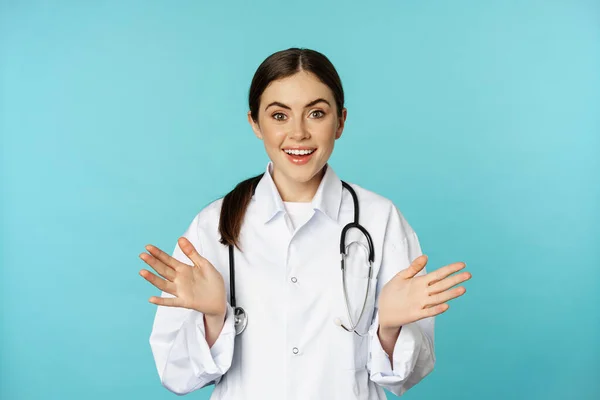 Happy and excited smiling young woman looking with excitement and surprised, standing in white lab coat and stethoscope, blue background — ストック写真