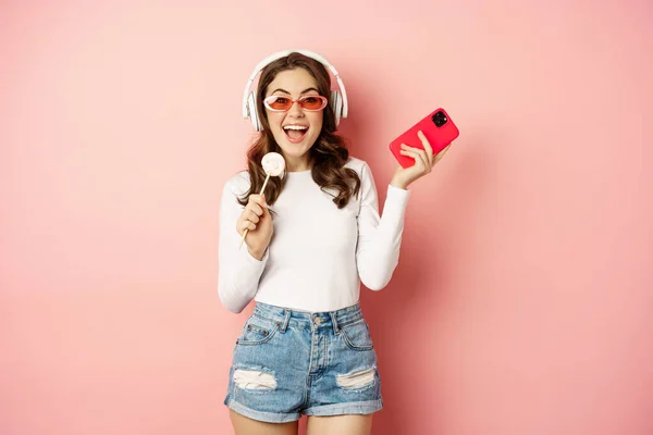 Letnia dziewczyna śmieje się, odziewanie lolipop i słuchanie muzyki w słuchawkach, taniec ze smartfonem na różowym tle — Zdjęcie stockowe