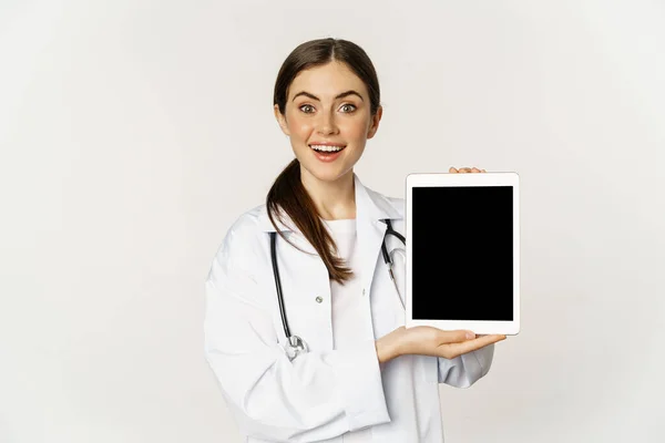 Imagen de la mujer doctora, trabajadora de la salud mostrando el sitio web médico en línea, la pantalla digital de la tableta y sonriendo, de pie en bata blanca sobre fondo blanco — Foto de Stock