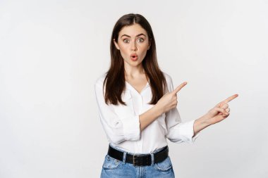 Amazed young woman pointing fingers down, showing announcement or logo banner, looking surprised and intrigued, standing over white background