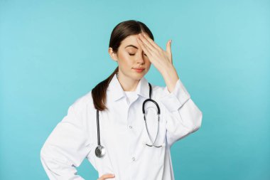 Portrait of annoyed, tired woman doctor, facepalm, roll eyes frustrated, bothered by smth stupid, standing in white coat over torquoise background