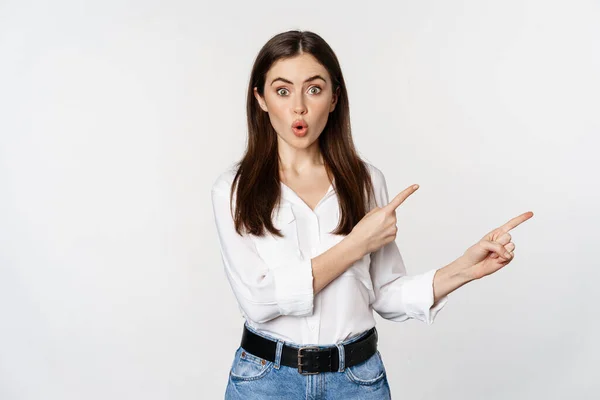 Amazed young woman pointing fingers down, showing announcement or logo banner, looking surprised and intrigued, standing over white background — Stock fotografie